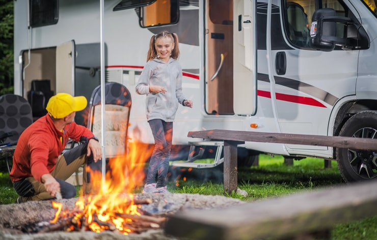 father and daughter on a road trip enjoy a fire in front of their class B RV at a wooded RV park